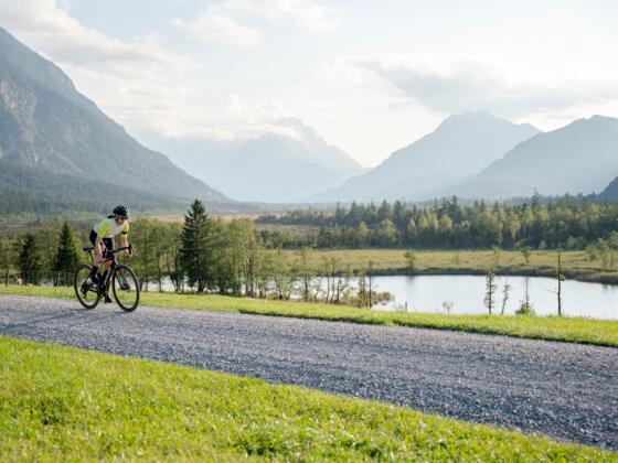Eine Gravelbikerin fährt über einen Feldweg in der malerischen Zugspitz Region. Im Hintergrund sind Seen, Wiesen und Berge.