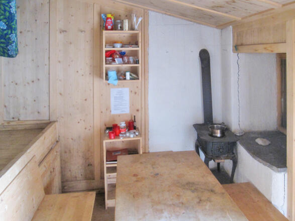 Innenraum der Linderhütte mit Schlafplatz, Kochstelle (Ofen) und Sitzgelegenheit