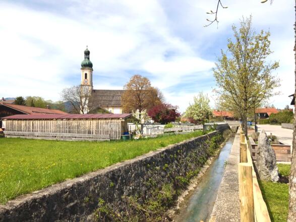 Entlang des Lenggrieser Dorfbaches finden sich die Kräuterhochbeete des Kräuterpfads mit Blick auf die Pfarrkirche