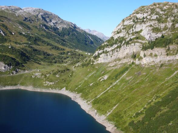 Der Felsensteig (rechte Bildhälfte) verläuft hoch über dem Formarinsee dicht unter den hellgrauen Steilwänden. Er benutzt die nur mäßig steile Böschung des darunter liegenden Gesteins